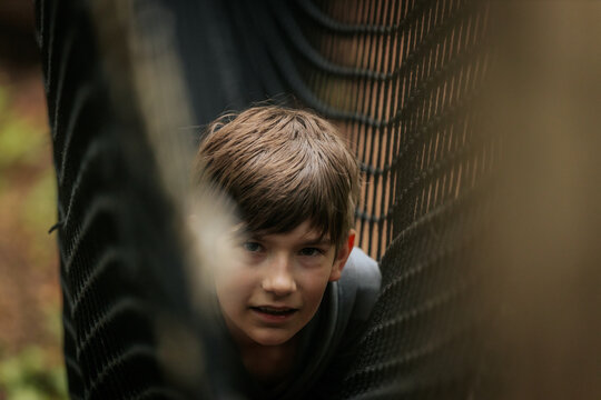Smiling boy in a gray hoodie gives a thumbs-up while lying inside a black rope net tunnel at an outdoor play area.
