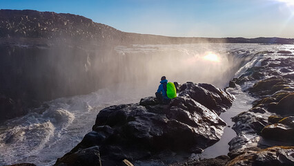 Lone hiker with a bright green backpack sits peacefully on a wet rock, contemplating powerful Selfoss waterfall in Iceland. The scene is enveloped in mist, backlit by low sun, creating a moody feel