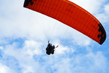 A pilot under an orange paraglider against a cloudy sky.