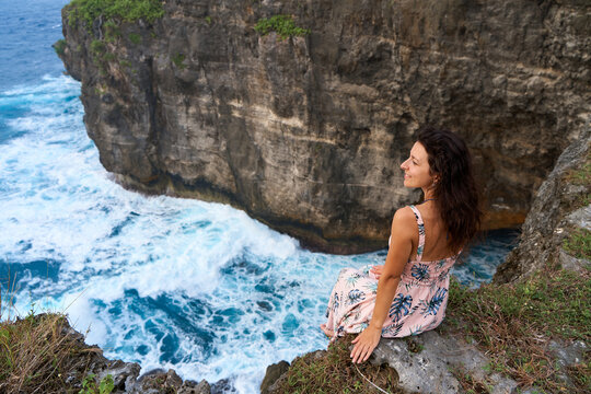 A beautiful woman in a pink dress sits on a cliff above the ocean on the island of Nusa Penida. Devil's Billabong an incredibly wonderful lagoon with splashes from the waves.