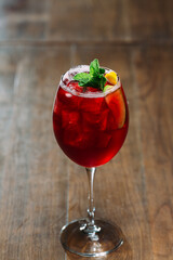 Red cocktail with ice, mint, and fruit garnish on wooden table