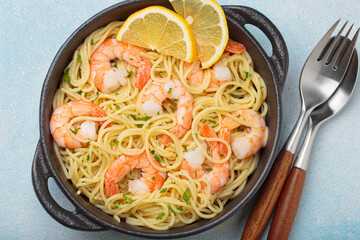 Shrimps pasta spaghetti served in a black cast iron skillet garnished with fresh lemon slices and parsley top view on light blue rustic background