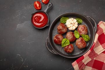 Fried meatballs in a cast iron pan, surrounded by basil, aromatic garlic, and tomato sauce on black background, healthy home cooking, space for text