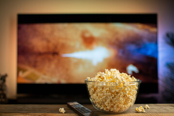 A glass bowl of popcorn and remote control in the background the TV works. Evening cozy watching a movie or TV series at home