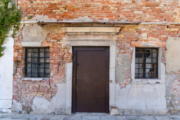 Old weathered brick wall with rustic door and windows. Aged brick and plaster facade with a dark wooden door and barred windows, showing signs of decay and historic charm.