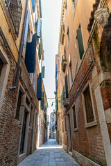 Fototapeta premium Narrow cobblestone street with open shutters in Venice. Charming narrow street in Venice with brick buildings, open wooden shutters, balconies, and soft light illuminating the alleyway.