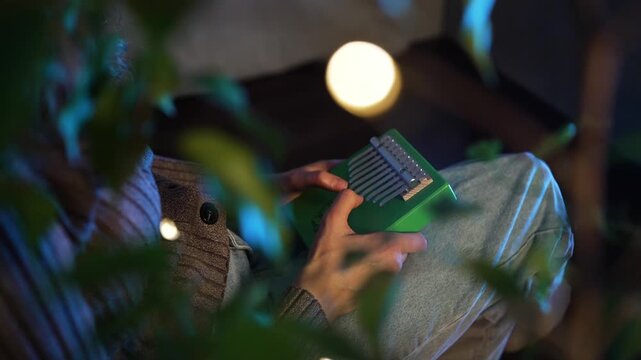 Young woman playing green kalimba, sitting comfortably in warm lighting surrounded by houseplants, creating peaceful musical moment in home environment