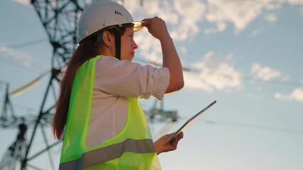 Woman engineer, power engineer in helmet checks power line using computer tablet online. Electrician in outdoors. Electric lines of high voltage at sunset. Distribution and supply of electricity