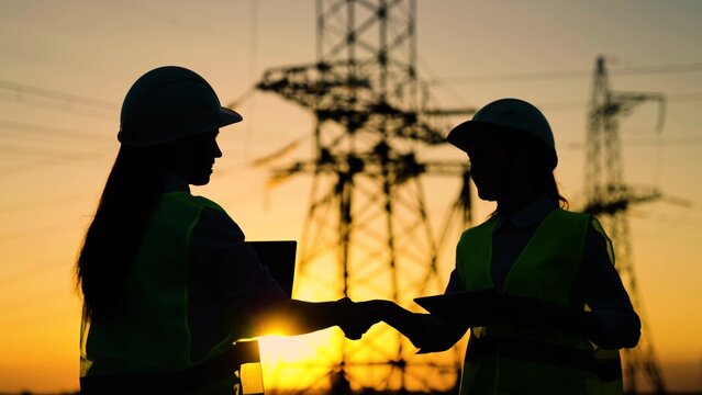 Colleagues women shake hands. Teamwork of power engineers in protective helmets, maintenance of power lines in outdoors. Two construction engineers work together on an electrical transmission line.