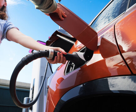 Close up of a woman and child connecting a charging cable to an electric car at a public station under a blue sky, symbolizing clean energy, sustainable travel and modern lifestyle