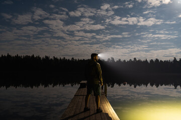 Adventurous man with a headlamp standing on a wooden dock by a calm lake, enjoying the beautiful moonlit night sky. © Dmitri