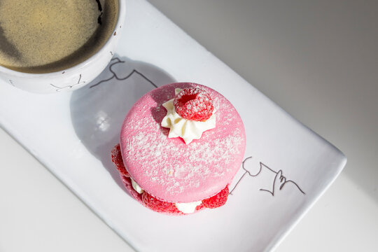 Elegant pink raspberry macaron with cream filling and fresh berries served alongside coffee on white porcelain plate. Perfect French pastry dessert presentation.