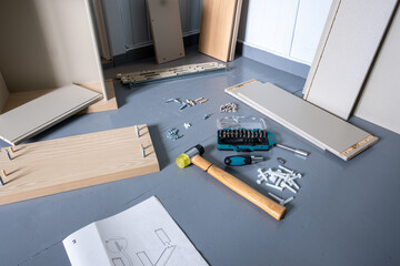 Furniture assembly in progress with wooden panels, tools, screws, dowels, and an instruction manual visible on the floor in a partially built room