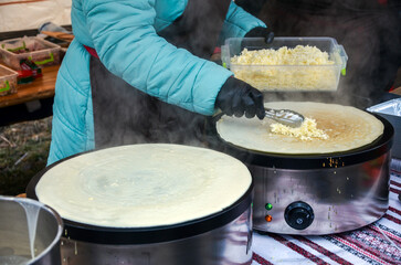 Person wearing a light blue jacket and black gloves is making crepes on two large, round griddles sprinkling cheese into one of the crepes