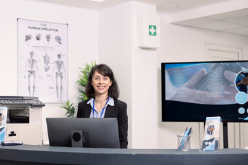 Hospital front desk with friendly caucasian female receptionist standing in clean, modern medical lobby. Professional clinic staff member ready to assist patients with registration.