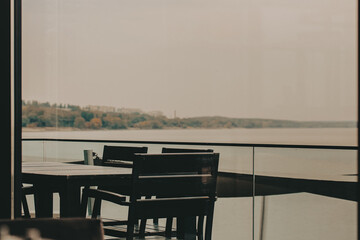 Outdoor dining area with a view of the lake on an overcast day. A dark table, chairs are arranged on a deck, with a serene lake view visible through the glass railing, creating a tranquil atmosphere.