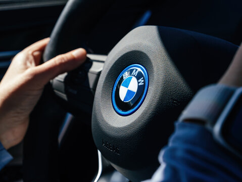 Baden, Germany - Sep 15, 2024: Detail of a BMW steering wheel logo with a driver's hand in frame, showcasing the blue and white emblem under sunlight inside a modern car interior