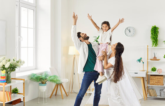 Happy family playing, father and mother holding excited daughter with hands raised up and laughter. Portrait of dad, mom and girl standing in hygge living room with tent at home. Love, game concept