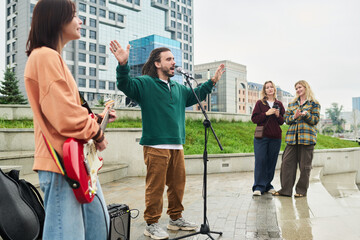 Caucasian young man singing into microphone with arms raised while Caucasian young woman playing electric guitar outdoors, two Caucasian young women standing nearby watching performance