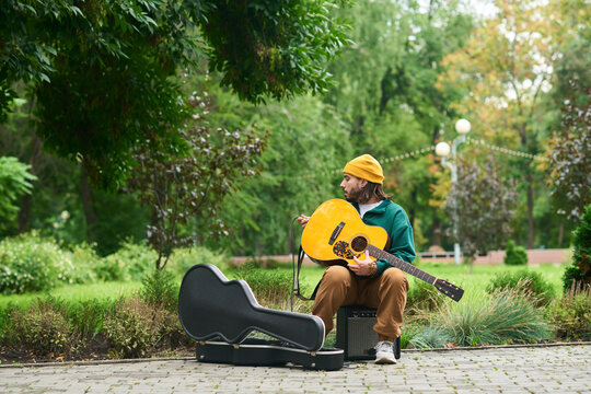 Young adult Caucasian man sitting outdoors tuning acoustic guitar, looking to side, guitar case open on ground, surrounded by green park vegetation, casual posture, facial hair visible