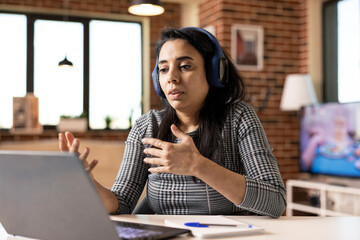 Indian female freelancer sits indoors with headphones, talking during video call with clients. Professional woman works remotely from modern brick wall apartment using laptop, having online discussion