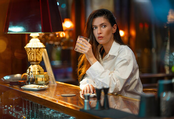 Attractive woman drinking alcohol at hotel bar with warm lighting and vintage decor. Beautiful young lady in white shirt and sitting calmly with thoughtful expression