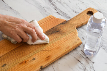 Closeup of a woman’s hand treating a wooden cutting board with mineral oil. Concept of kitchen care, wood maintenance, seasoning, and protection using natural household methods