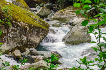 Fototapeta premium Mountain stream rushing through mossy boulders and rocks. Creating cascades of water. Surrounded by greenery, the fast-flowing river showcases the raw beauty and freshness of a natural landscape.