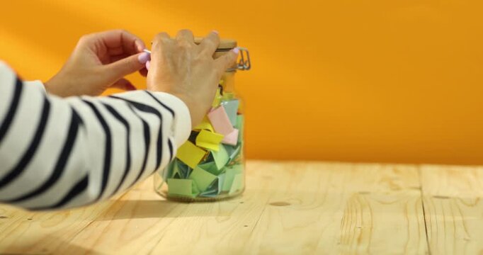 Woman folding yellow paper piece and putting it into glass jar at wooden table, closeup