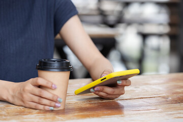 A person sits at a wooden table holding a takeaway coffee cup in one hand and a yellow smartphone in the other, symbolizing modern lifestyle, digital connection, and casual relaxation.