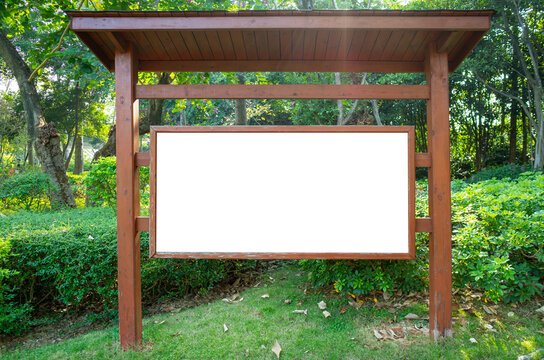 Wooden outdoor signboard with blank white display panel under a roofed structure, placed in a green public park. Mockup copy space for text, information, community announcement or advertisement.