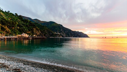 Peaceful sunset at a Mediterranean coast with sailboats, pebbled beach, lush hills, and a pier reflecting warm evening light.
