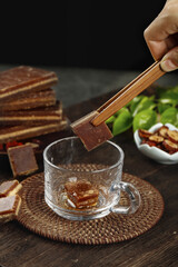 Hand stirring tea with large brown sugar block cube using wooden stick on rustic table with mint leaves