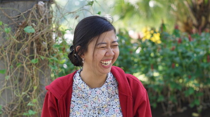 Portrait of a young Asian woman wearing a red jacket and floral blouse, standing outdoors with green plants and natural background, looking calm and confident
