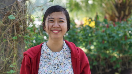 Portrait of a young Asian woman wearing a red jacket and floral blouse, standing outdoors with green plants and natural background, looking calm and confident