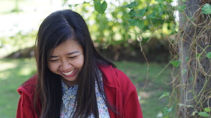 Portrait of a young Asian woman wearing a red jacket and floral blouse, standing outdoors with green plants and natural background, looking calm and confident