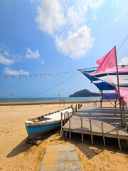 A small boat next to pier.A small boat with a light blue hull and a weathered, white top is pulled up onto a sandy beach, next to a wooden pier. 