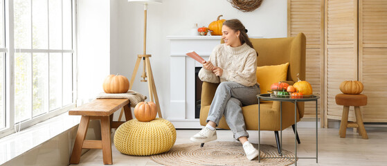 Beautiful young happy woman reading magazine and sitting on armchair in room decorated for autumn