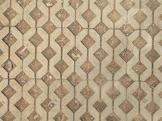 A close-up of interlocking concrete pavers creates a geometric, diamond-patterned texture. The earthy, neutral tones of the blocks are highlighted by scattered leaves and dirt