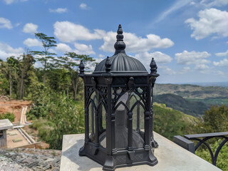 Obraz premium A black gothic-style lamp with intricate carvings sits on a concrete ledge, overlooking a panoramic view of a hilly green landscape and a vibrant blue sky.