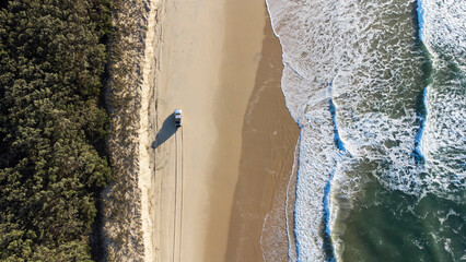 This aerial shot captures a serene beach scene with a 4WD vehicle parked on the sand. The ocean waves gently lap the shore, creating a foamy white border against the tan sand. 