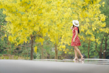Portrait young woman in red dress and white hat is relaxing on her vacation in a spring park with background of blooming yellow flowers of Golden Shower Tree, Khao Ngu Stone Park, Ratchaburi, Thailand