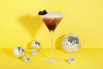 Glass with tasty cool blackberry lemonade and decorative disco balls on yellow background, closeup