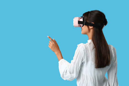 Young woman in modern VR glasses with mobile phone pointing at something on blue background, back view - Powered by Adobe
