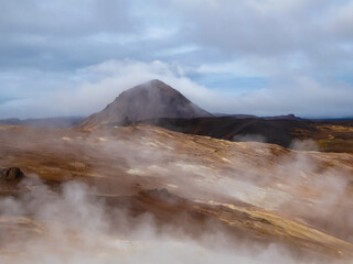 Billowing steam from geothermal vents rises over a barren, colorful landscape, partially obscuring the volcanic Námafjall mountain under a cloudy sky in the Hverir geothermal area of Iceland.