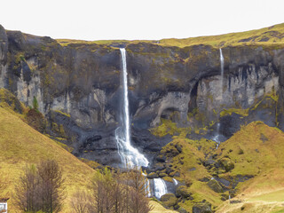 Several thin, delicate waterfalls cascade down a dark, moss-dappled cliff face in the Icelandic countryside, creating a complex and beautiful water feature set against grassy hillsides in autumn.