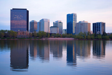 Naklejka premium Riverside cityscape at dusk with reflections of skyscrapers in calm water, soft evening light, tranquil urban scenery.
