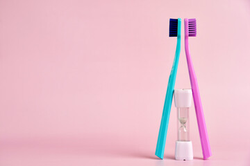 Two colorful toothbrushes and a small hourglass sit against a pastel pink background, illustrating the recommended brushing time