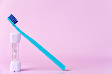 A turquoise toothbrush rests against a small white hourglass timer, set against a soft pink background, symbolizing the two-minute brushing time