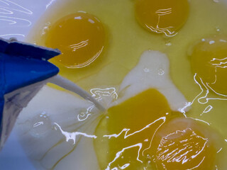 Raw eggs with yolks and whites in a white bowl, milk being poured from carton, close-up view of...